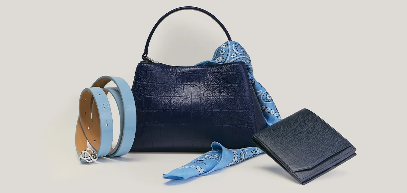 Bolso azul marino con pañuelo azul estampado, cinturón doble azul claro y beige, y cartera azul marino dispuestos sobre superficie beige.