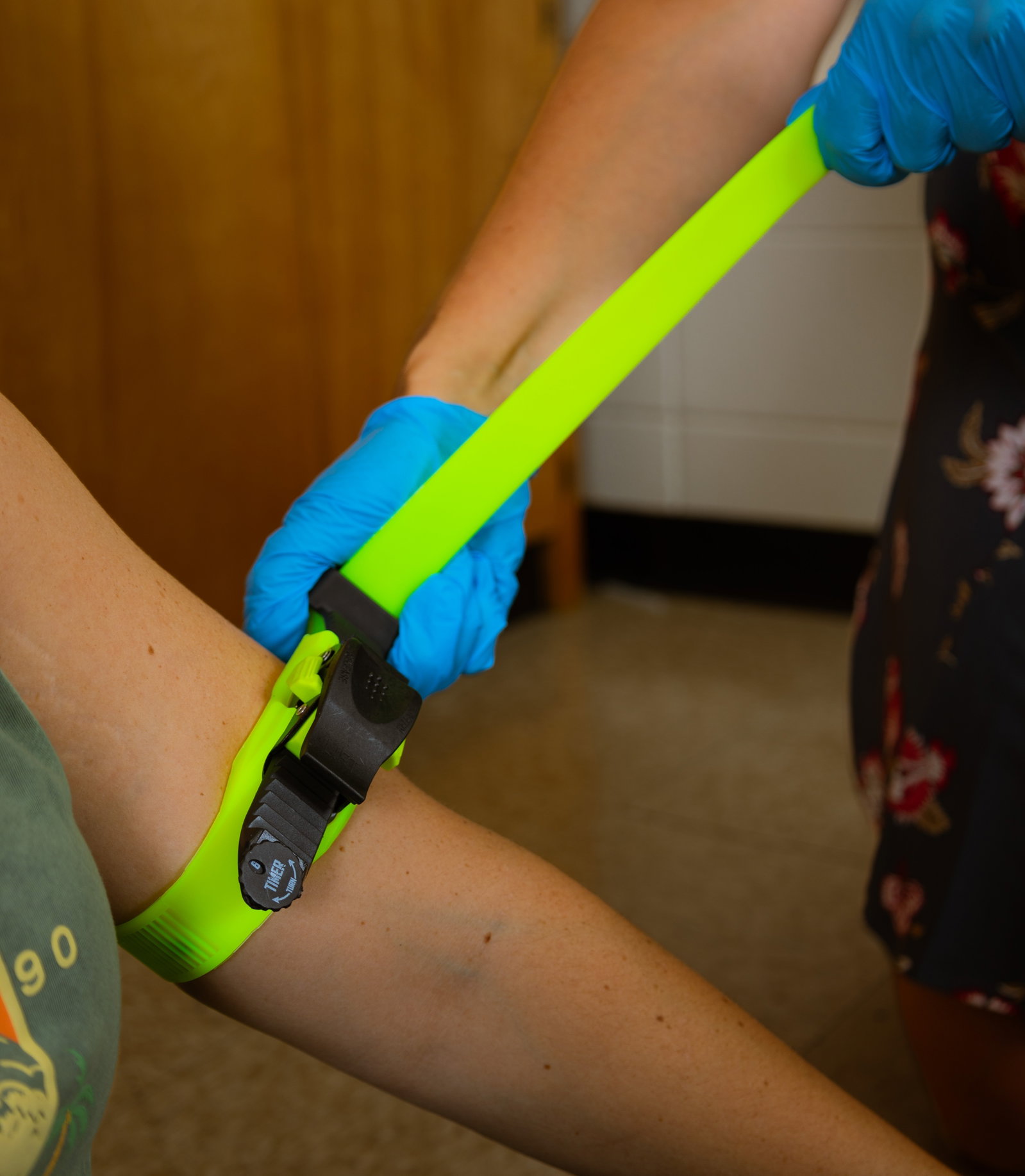 Gloved hands applying a bright lime ratcheting tourniquet to an upper arm; close-up, indoor.