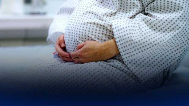 The torso of a pregnant woman wearing a hospital gown with her hands resting on her stomach.