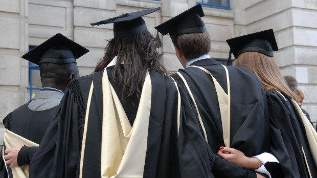 Four students dressed in graduation robes and mortarboards stand with their backs to the camera. They have their arms around one another.