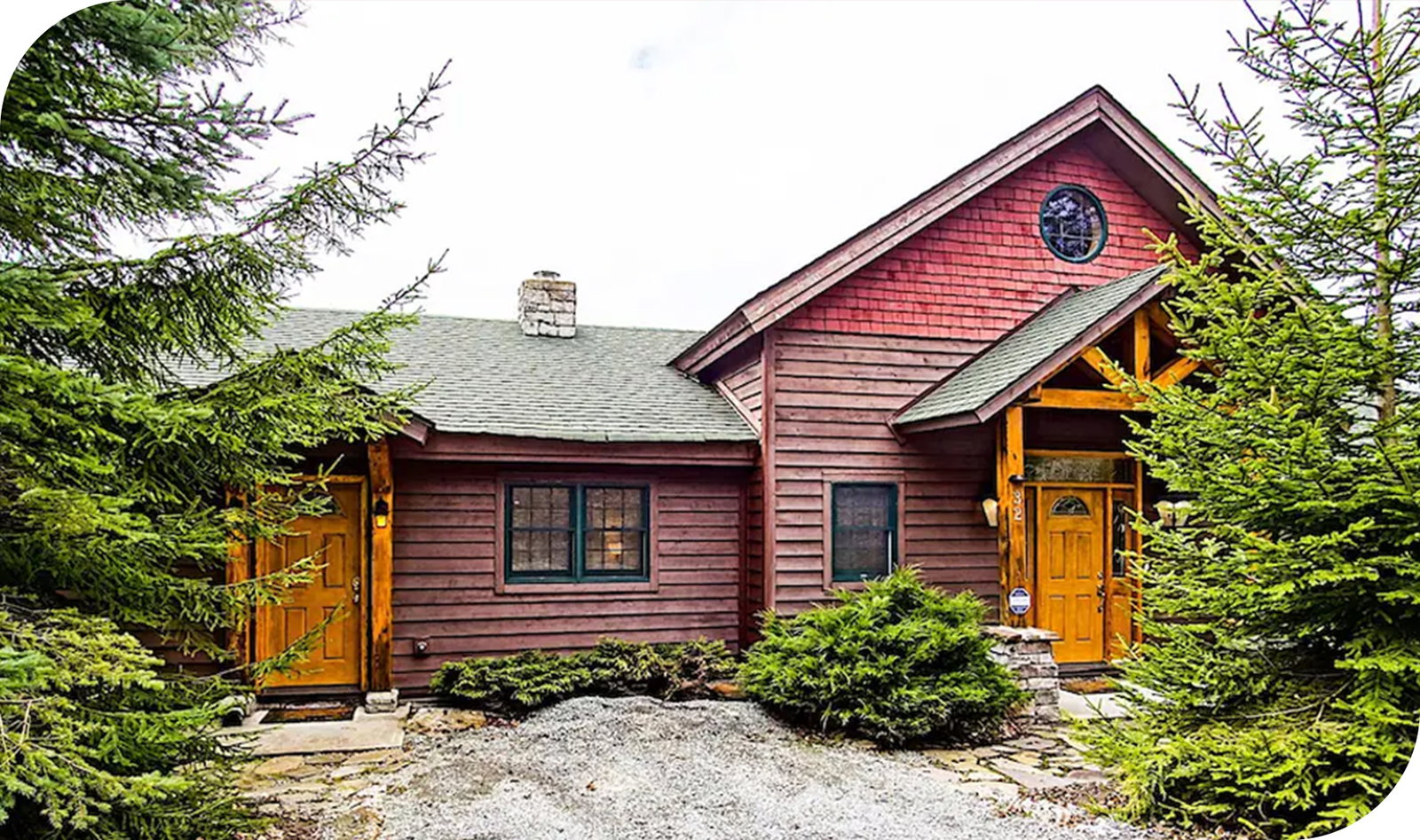 Rustic wood cabin surrounded by evergreen trees with dual front doors and gravel path.