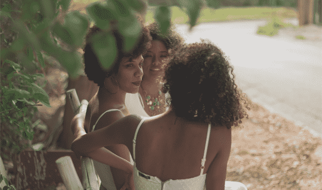 Three people with curly hair sit on a bench, engaged in conversation. They are outdoors, surrounded by greenery.