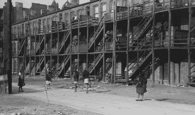 Children play in front of a row of old brick apartment buildings with wooden staircases and balconies in an urban area. Debris is scattered on the ground.