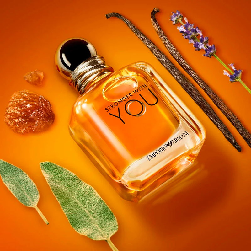 Armani Stronger With You EDT fragrance bottle displayed with vanilla pods, chestnut, and sage leaves on a radiant golden-orange backdrop.