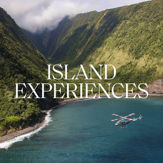 Helicopter flying past a lush mountain valley with the ocean below. Text overlay: Island Experiences.