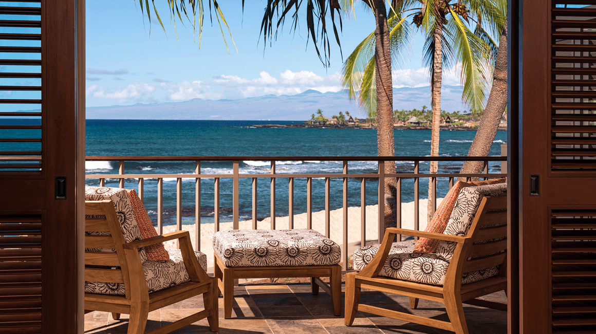 Two cushioned outdoor chairs and an ottoman on a balcony overlooking the ocean.