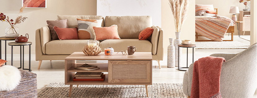 Helles Wohnzimmer mit beigem Sofa, mehreren Kissen in warmen Tönen und einem hellen Holztisch in der Mitte. Auf dem Tisch stehen eine dekorative Schale, Kerzen und eine Tasse. Links und rechts vom Sofa befinden sich kleine runde Beistelltische mit Vasen und Trockenblumen. Im Hintergrund ist ein Bett mit gestreifter Decke zu sehen. Ein grauer Sessel mit rostrotem Kissen steht rechts im Vordergrund. Der Raum wirkt freundlich und modern eingerichtet.
