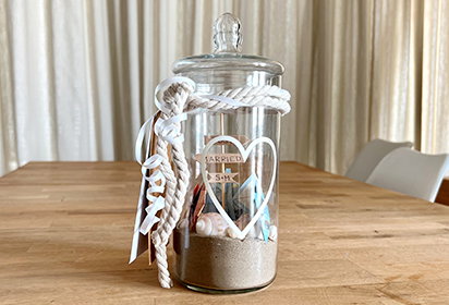Auf einem großen Holztisch steht ein hohes Glas mit einem Seil umwickelt. Auf dem Glas ist ein Herz gedruckt. Im Glas befindet sich Sand, Muscheln und weitere maritime Dekoelemente. Rechts steht der Text ‚MARITIME SCHATZKISTE‘ mit einem Button ‚Zum DIY‘.