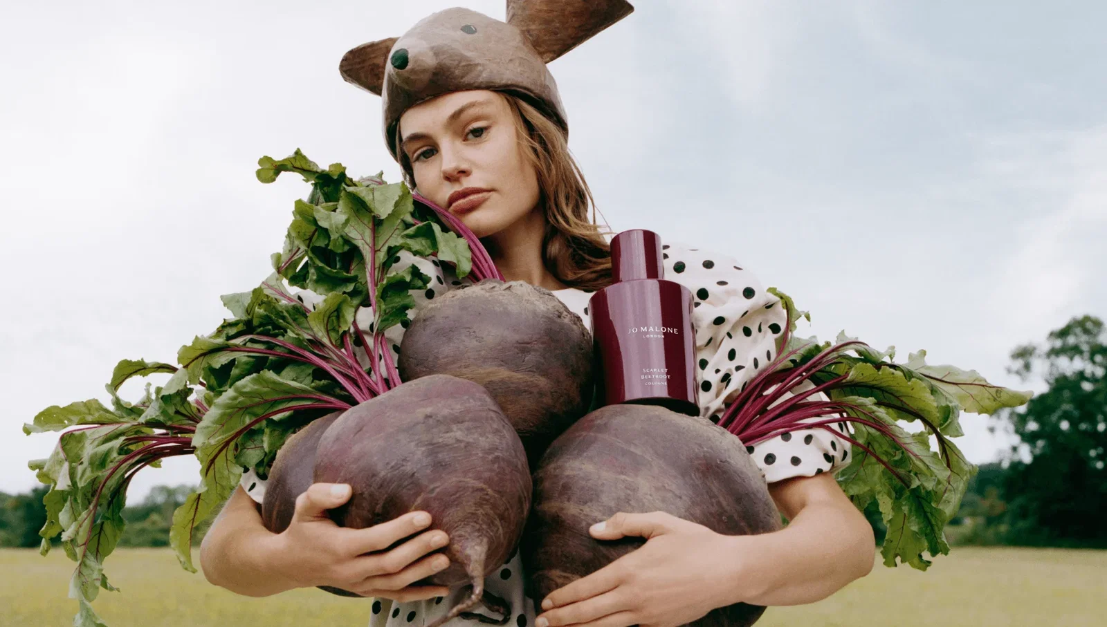 Woman holding four giant beetroots and a bottle of Jo Malone Scarlet Beetroot Cologne, wearing an animal hat and polka-dot clothing in the countryside.
