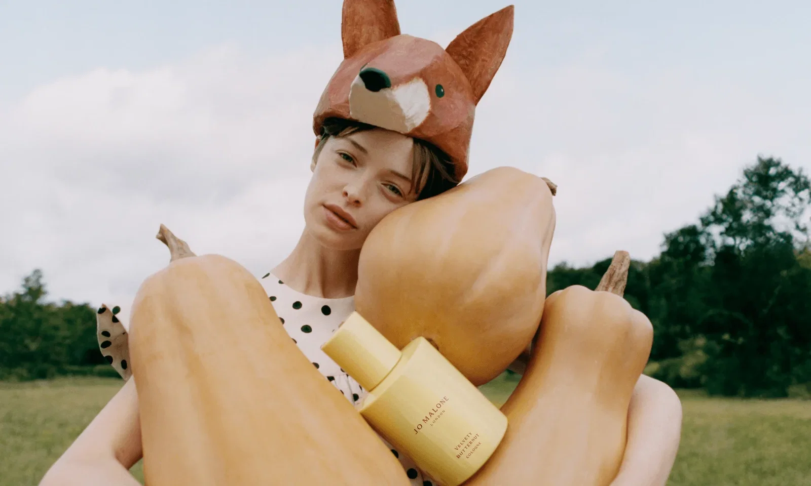 Woman holding three giant butternut squashes and a bottle of Jo Malone Velvety Butternut Cologne, wearing a fox hat and polka-dot clothing in the countryside.