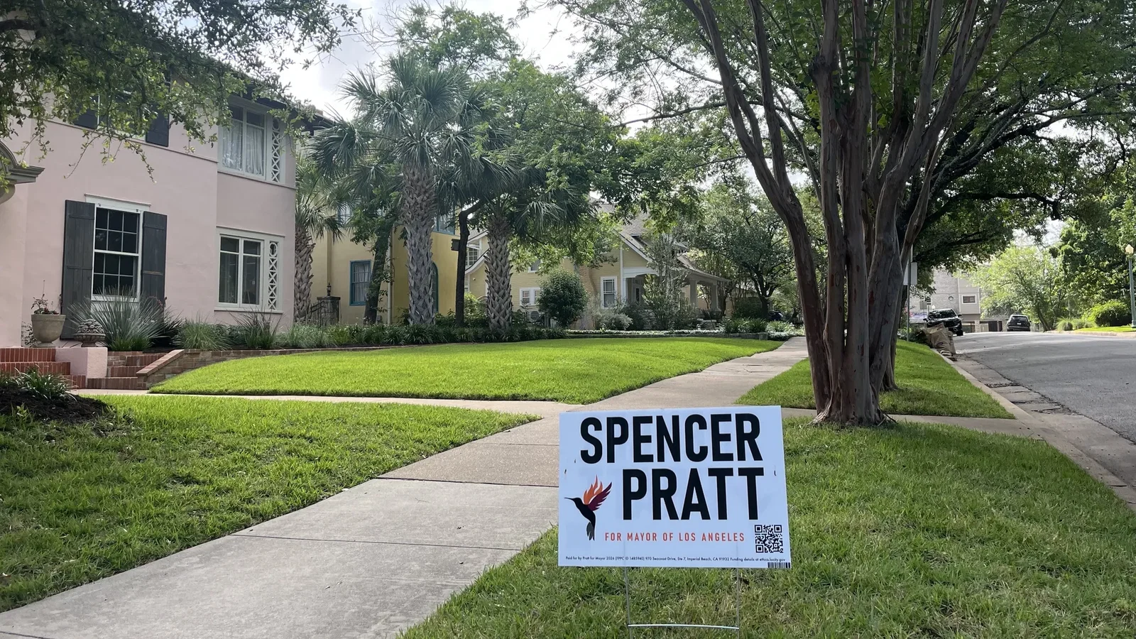 Suburban street scene with pastel pink house, green lawn, and tall trees including palms. A blue and white campaign sign in the foreground reads SPENCER PRATT for mayor.