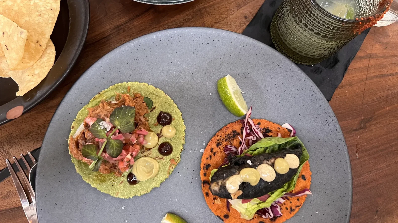 Top-down view of a wooden table with colorful tostadas on orange and green tortillas, a lime wedge, chips, a guacamole bowl, and a textured glass with a drink on the right.