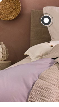 Ein Bett steht vor einer lila Wand. Auf dem Bett befindet sich lila- und rosafarbige Bettwäsche sowie ein gestreiftes Dekokissen und eine Tagesdecke. Links und rechts neben dem Bett steht jeweils ein Steintisch. Links steht eine kleine Vase darauf und rechts eine große Vase mit Blumen. 