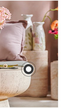 Auf einem Steintisch steht eine Vase mit Blumen darin. Im Hintergrund ist ein Sofa zu sehen mit einem rosafarbigen und gestreiften Kissen sowie weiteren Steintischen mit Blumenvasen.