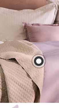 Ein Bett steht vor einer lila Wand. Auf dem Bett befindet sich lila- und rosafarbige Bettwäsche sowie ein gestreiftes Dekokissen und eine Tagesdecke. Links und rechts neben dem Bett steht jeweils ein Steintisch. Links steht eine kleine Vase darauf und rechts eine große Vase mit Blumen. 