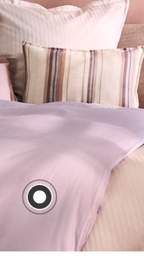 Ein Bett steht vor einer lila Wand. Auf dem Bett befindet sich lila- und rosafarbige Bettwäsche sowie ein gestreiftes Dekokissen und eine Tagesdecke. Links und rechts neben dem Bett steht jeweils ein Steintisch. Links steht eine kleine Vase darauf und rechts eine große Vase mit Blumen. 