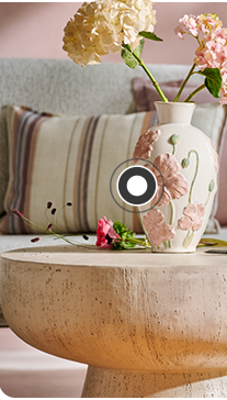Auf einem Steintisch steht eine Vase mit Blumen darin. Im Hintergrund ist ein Sofa zu sehen mit einem rosafarbigen und gestreiften Kissen sowie weiteren Steintischen mit Blumenvasen.