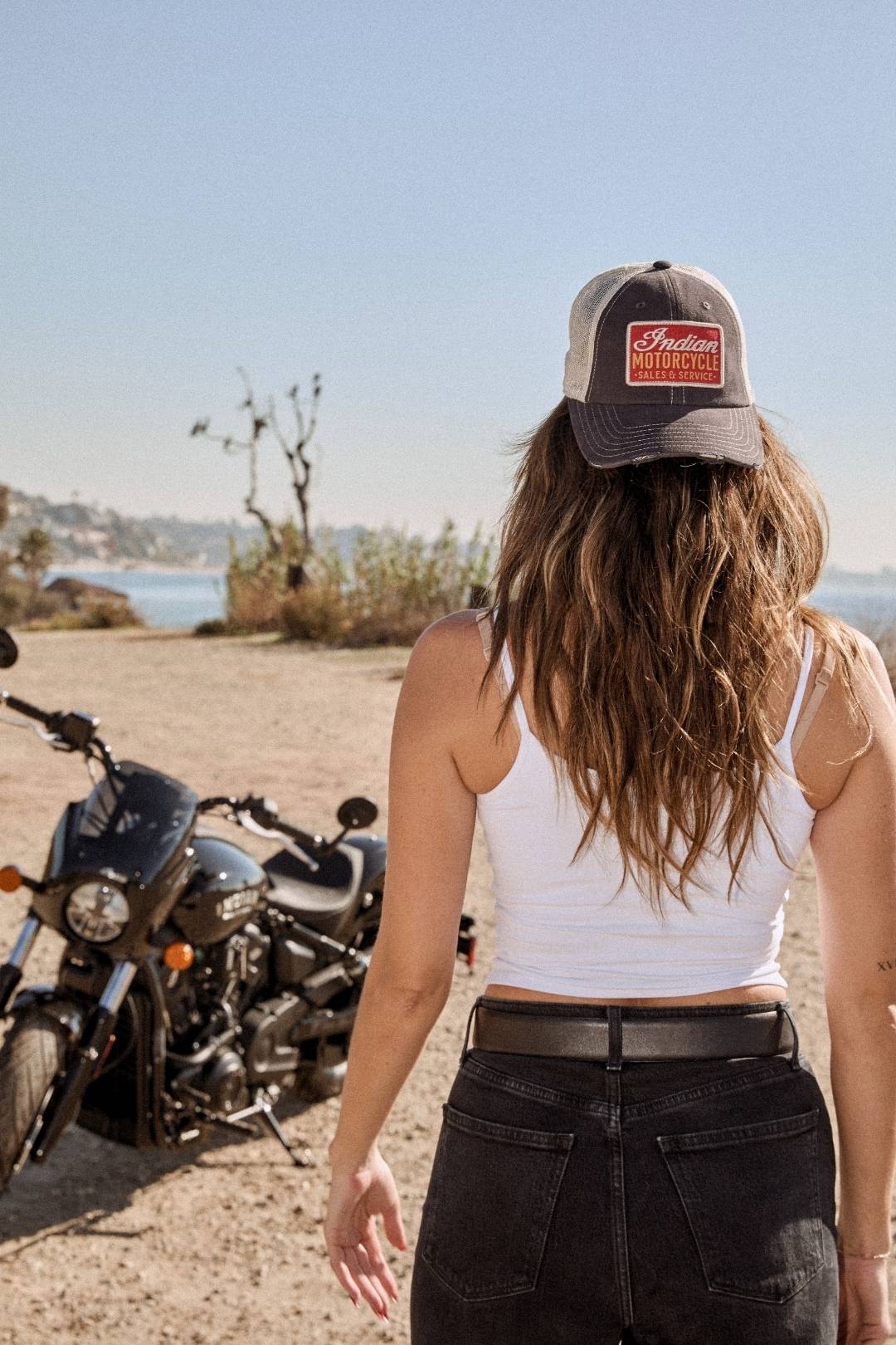 A woman seen from behind wears a white tank top, black jeans, and a red and brown Indian Motorcycle Orville hat. She is standing next to a parked black motorcycle on a dirt road overlooking the ocean.