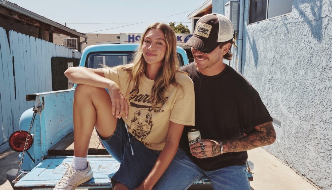 A smiling man and woman sitting together on the open tailgate of a vintage light blue pickup truck. The man is wearing a dark brown and tan Garage Beer Orville hat, while the woman wears a Garage Beer RL Pigment tee.
