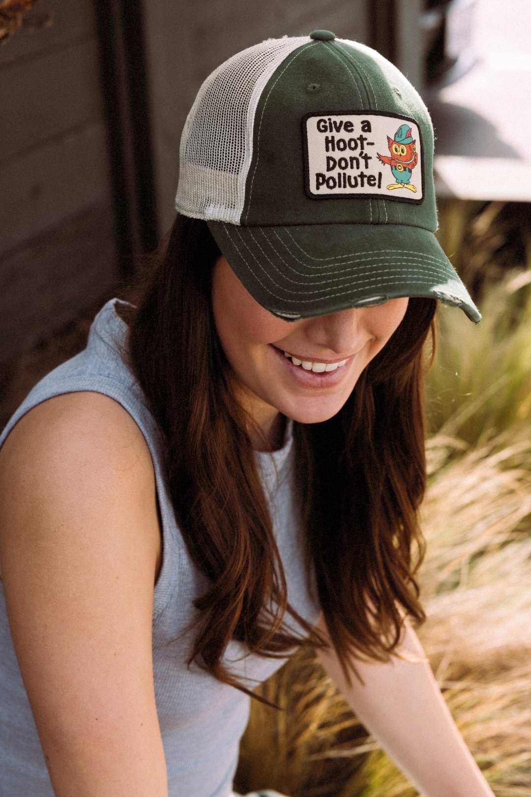 A close-up of a smiling woman looking downward, wearing a green mesh Orville trucker hat with a vintage Woodsy Owl 'Give a Hoot - Don't Pollute!' patch.