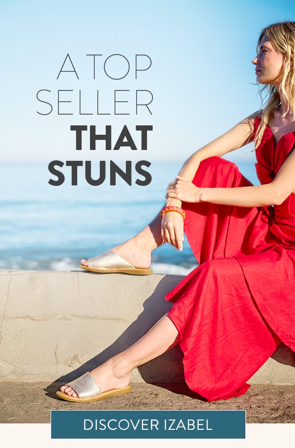 A Top Seller That Stuns. A woman sitting by the ocean wearing the Izabel flat slide sandal in metallic.