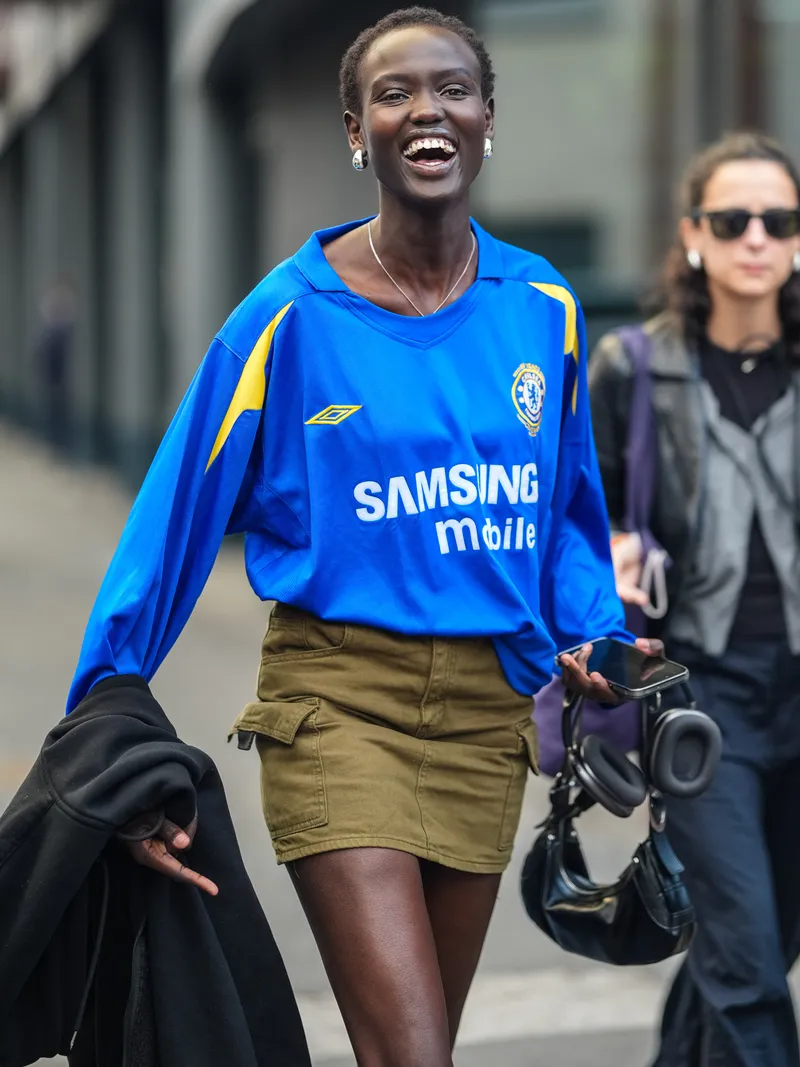how to style a soccer jersey MILAN, ITALY - SEPTEMBER 20: A model wears silver chrome earrings, silver necklace, bright blue oversized graphic print sports shirt, dark green olive denim jean mini skirt, shiny black leather bag, black hoodie, outside Tod's, during the Milan Fashion Week Spring/Summer 2024-2025 on September 20, 2024 in Milan, Italy. (Photo by Edward Berthelot/Getty Images)