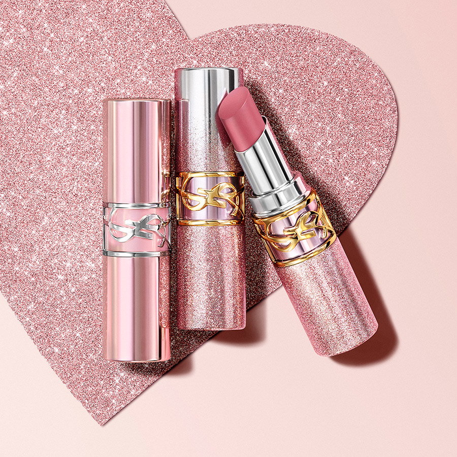Three tubes of YSL LOVESHINE CANDY GLAZE lipstick in shimmering silver and glittery rose gold packaging, arranged over a sparkling pink glitter heart background.