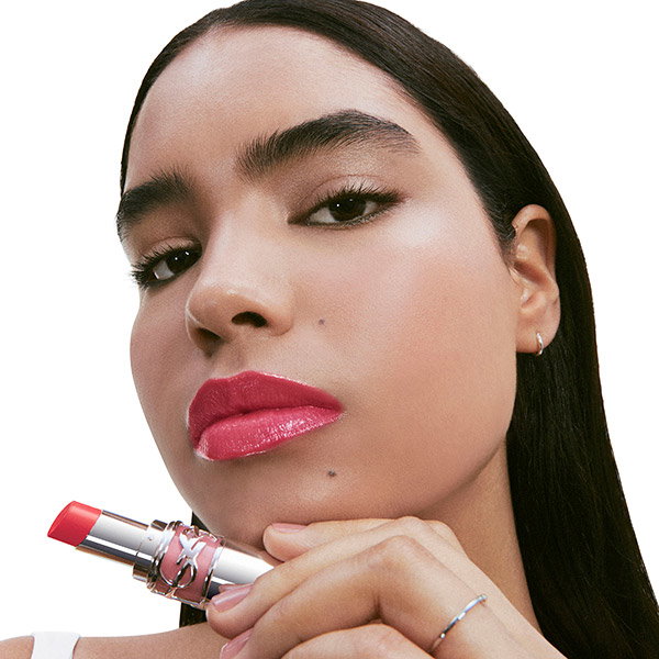 Close-up of a model with a BOLD magenta lip, holding a YSL LOVESHINE lipstick tube to her chin. She has a powerful and unapologetic expression.