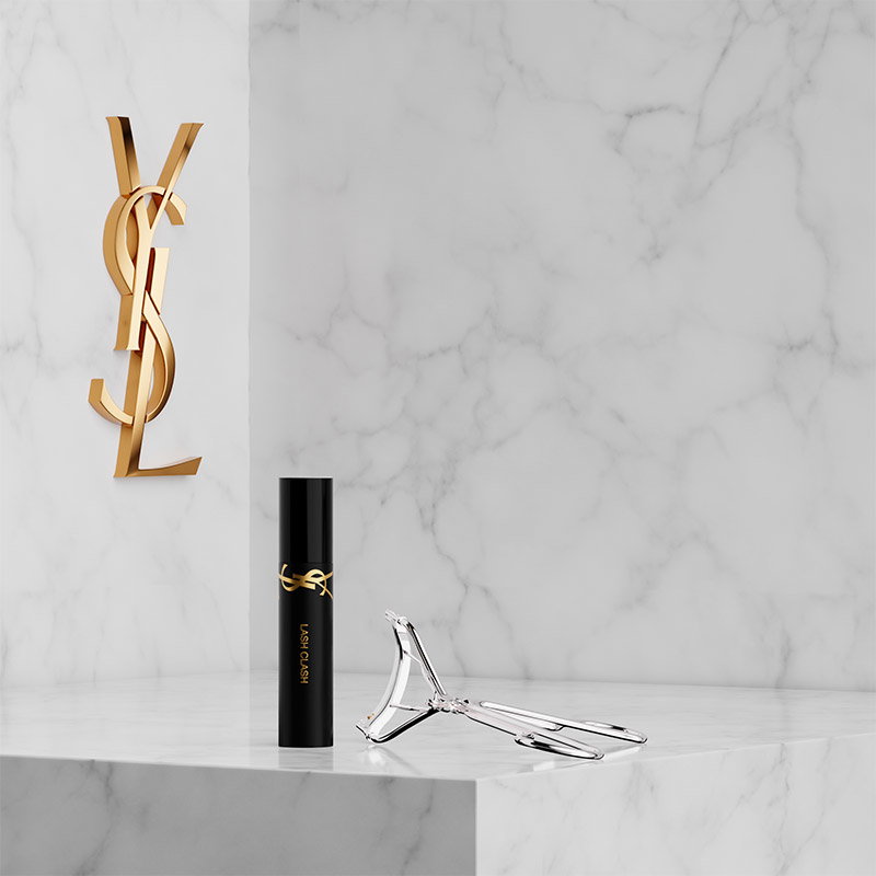 A MINI LASH CLASH MASCARA and a silver YSL EYELASH CURLER placed on a white marble pedestal next to a gold YSL Cassandre logo on a marble wall.