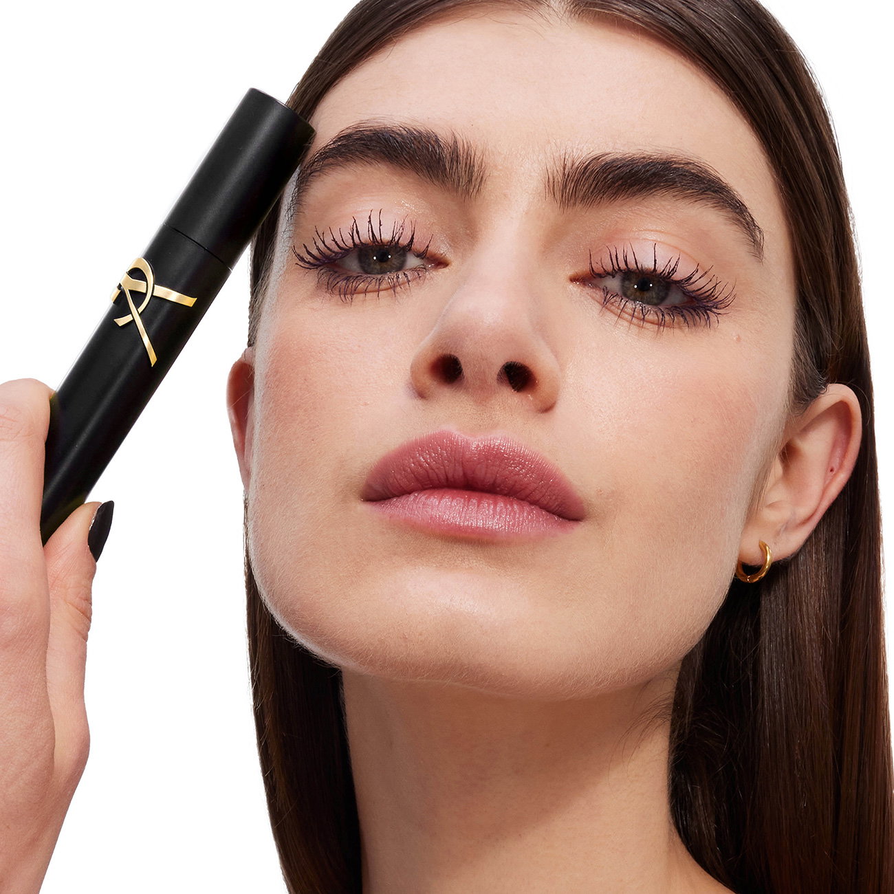 Close-up of a model with a FLAWLESS complexion and natural makeup, holding a YSL LASH CLASH mascara tube to her forehead. She has bold brows and a soft pink lip.