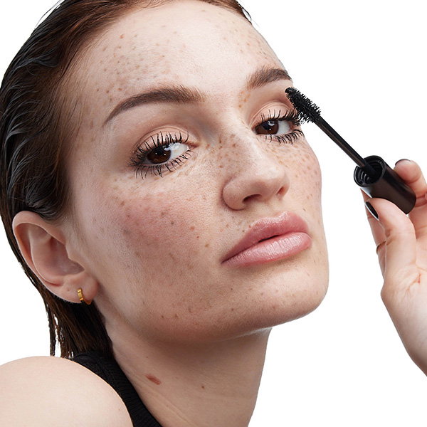 Close-up of a model with freckles applying black mascara to her upper lashes with a YSL LASH CLASH mascara wand. Her look is natural and radiant.