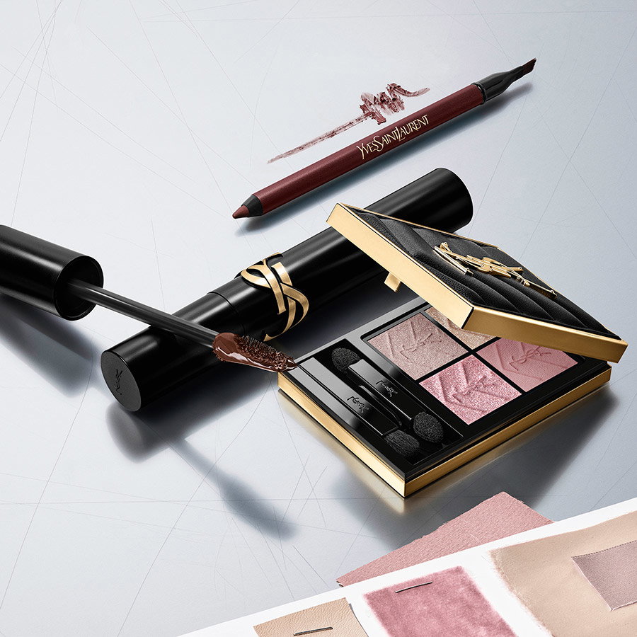 A flat-lay composition featuring a YSL COUTURE CLUTCH eyeshadow palette in pink and purple shades, a black eyeliner pencil with a purple smudge, and a black mascara tube with a gold YSL logo.