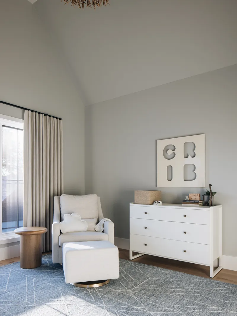 A glider, foot stool, end table and dresser sit in the corner of a room with minimal decor, an area rug, a window and vaulted ceilings