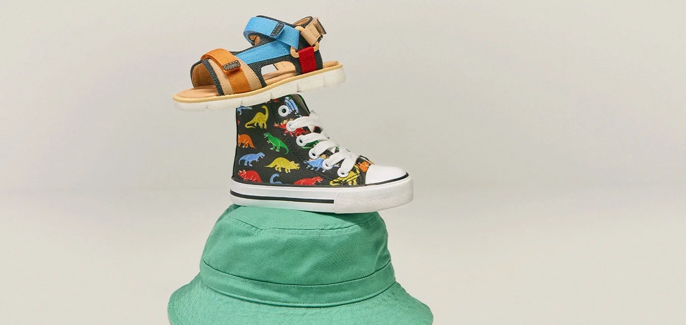 Gestapelde kinderaccessoires: groene emmerhoed, zwarte hoge sneaker met kleurrijke dinosaurussen, en blauwe en oranje klittenbandsandaal bovenop.