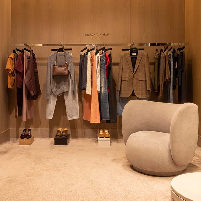 Brown Thomas fitting room with clothes on racks, a plush beige chair, and heeled shoes on display.