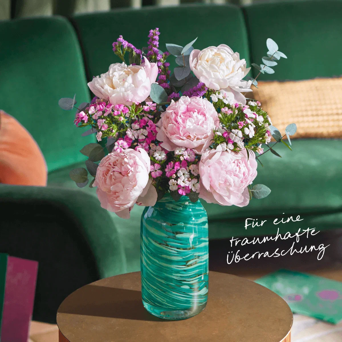 Ein Bouquet aus rosa Pfingstrosen, lila Meerlavendel und Eukalyptus steht in einer grün-blau gemusterten Glasvase. Daneben steht der Text: ‘Für eine traumhafte Überraschung’. Im Hintergrund ist ein grünes Sofa zu sehen.