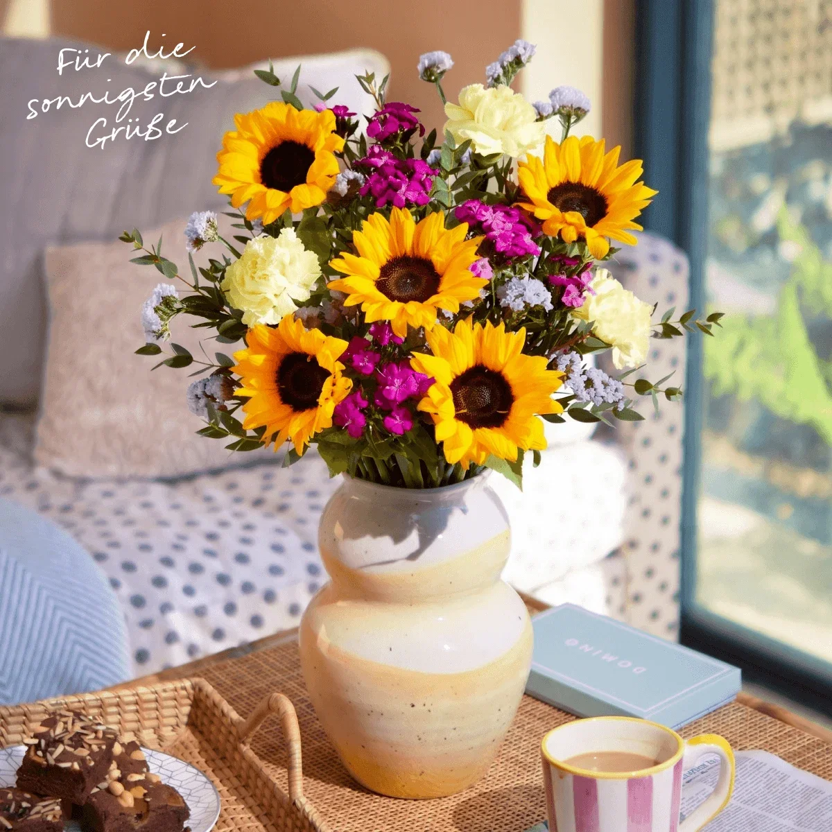 Eine gelb-weiß geschwungene Keramikvase ist mit Sonnenblumen, Nelken und pinken Bartnelken gefüllt. Oben links steht der Text: ‘Für die sonnigsten Grüße’. Sie steht auf einem Flechttisch neben Brownies und Kaffee vor einem Sofa im Hintergrund.