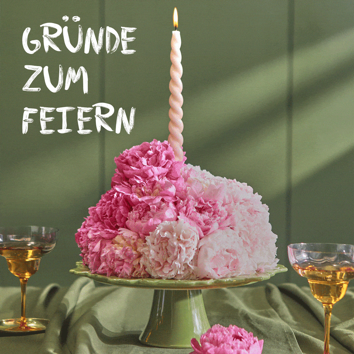 Ein grüner Tortenständer präsentiert ein rundes Gesteck aus pinken und hellrosa Pfingstrosen, gekrönt von einer brennenden, gedrehten Kerze. Links oben steht der Text „GRÜNDE ZUM FEIERN“. Champagnergläser rahmen das festliche Arrangement ein.