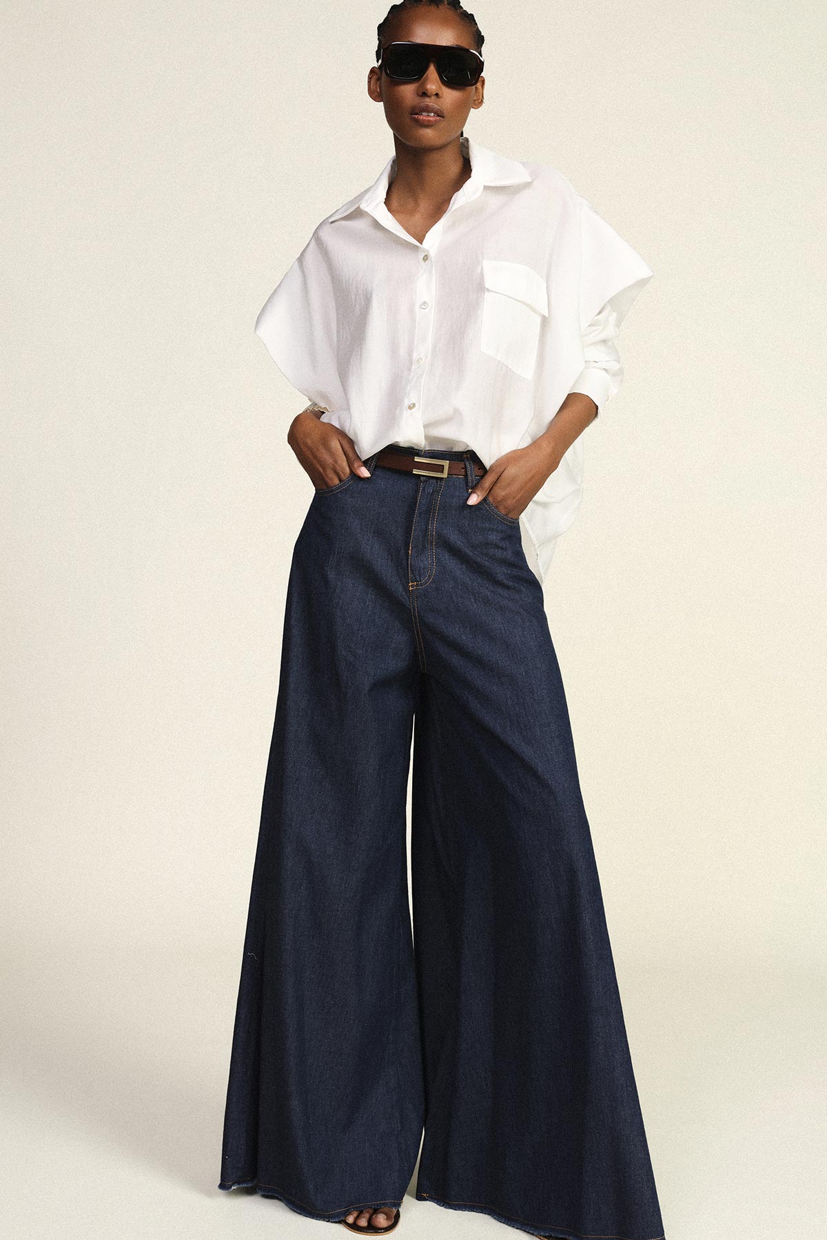 Woman modelling oversized white shirt and high-waisted wide-leg dark denim jeans with belt and sunglasses, hands in pockets