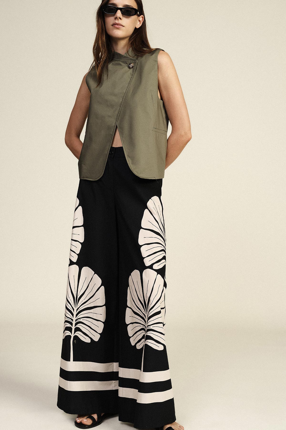 Woman wearing sleeveless olive-green jacket, black wide-leg trousers with large white leaf print and sunglasses