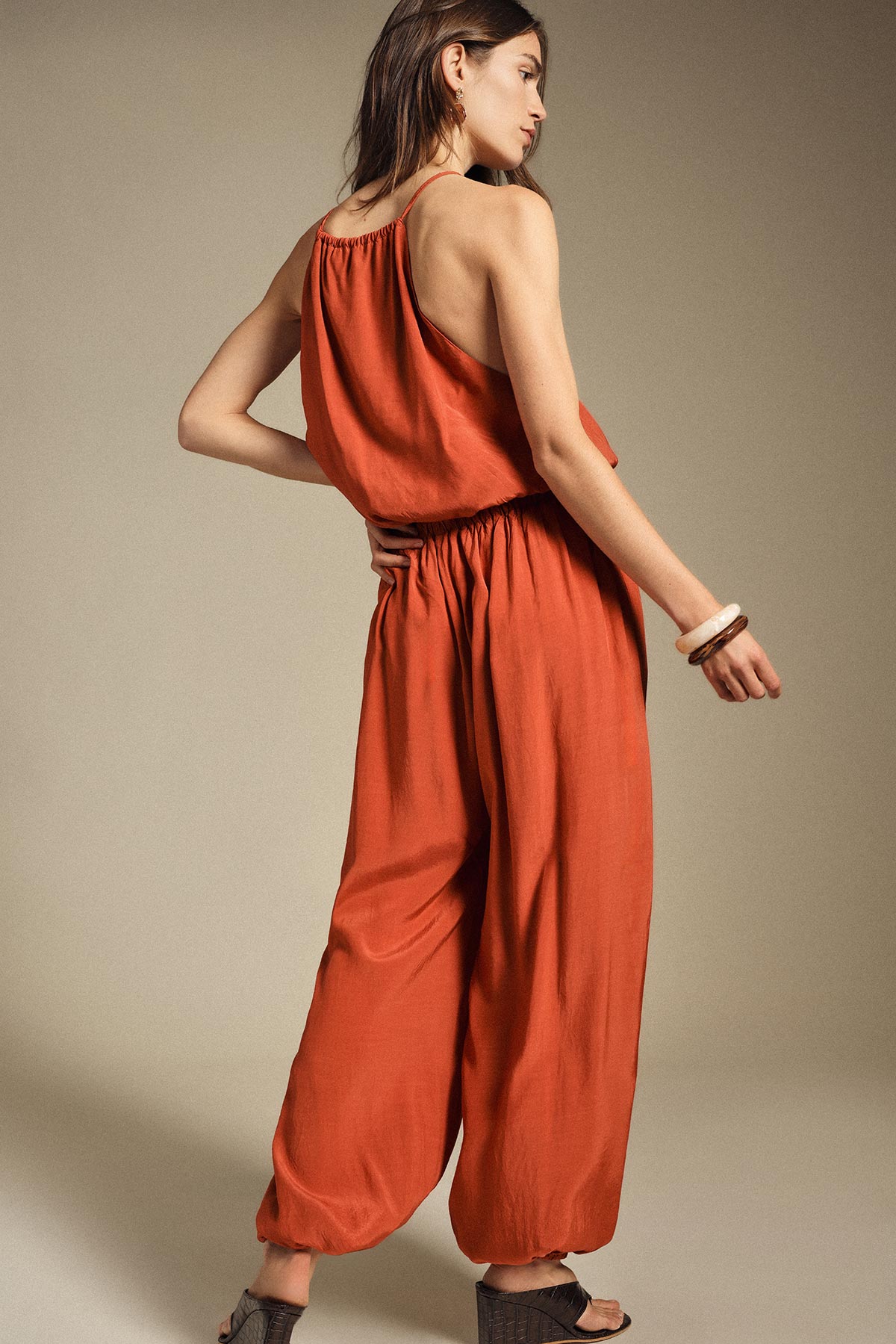 Woman modelling rust-coloured sleeveless wide-leg jumpsuit, back view, wearing wooden bangles and sandals on neutral background