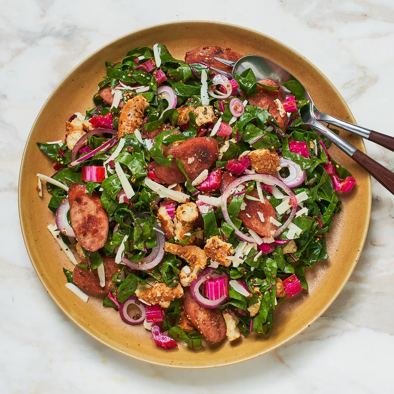 A salad with ribbons of Swiss chard, sliced sausage, onions, and more in a large bowl with salad servers.