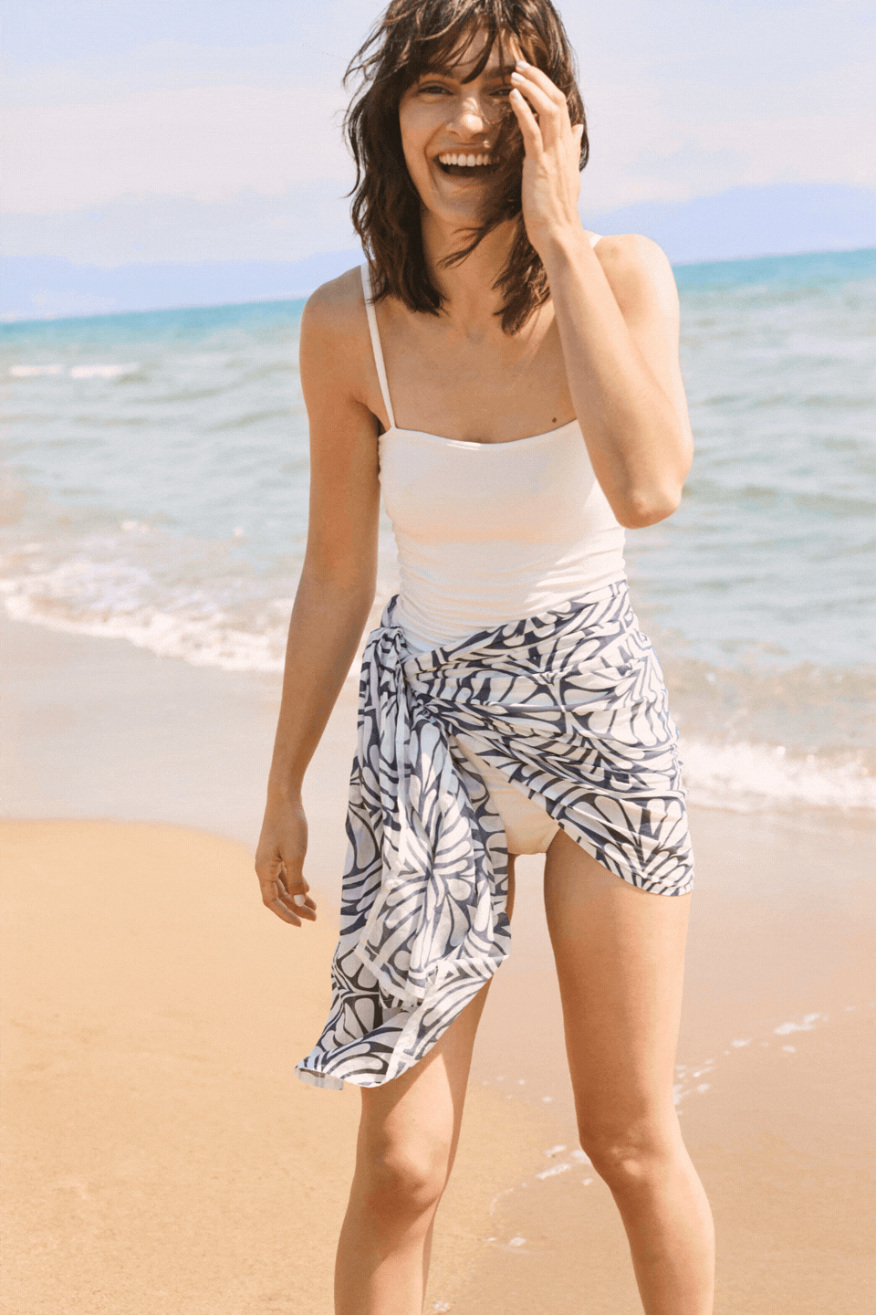 Smiling woman standing by the shoreline in a white swimsuit and patterned beach wrap, with soft waves and a bright coastal backdrop behind her.