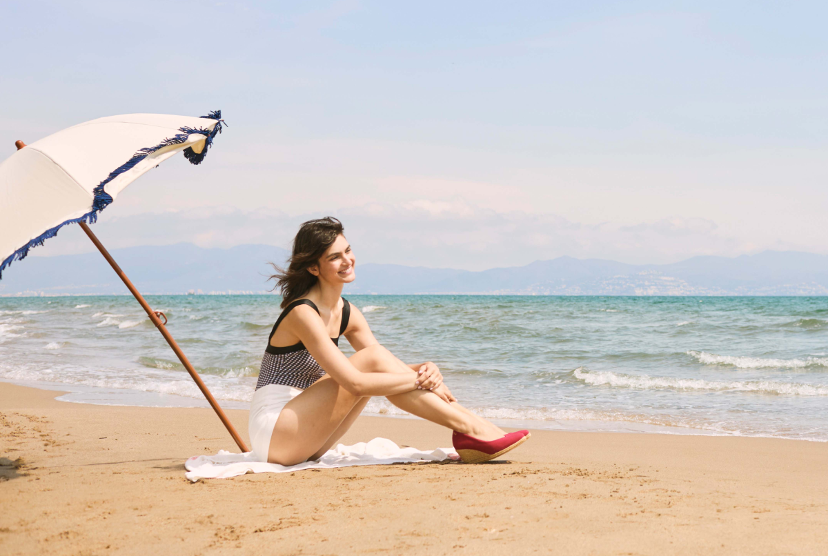 Woman sitting on a sandy beach beneath a fringed cream parasol, wearing red Viscata espadrilles while looking out toward the sea.