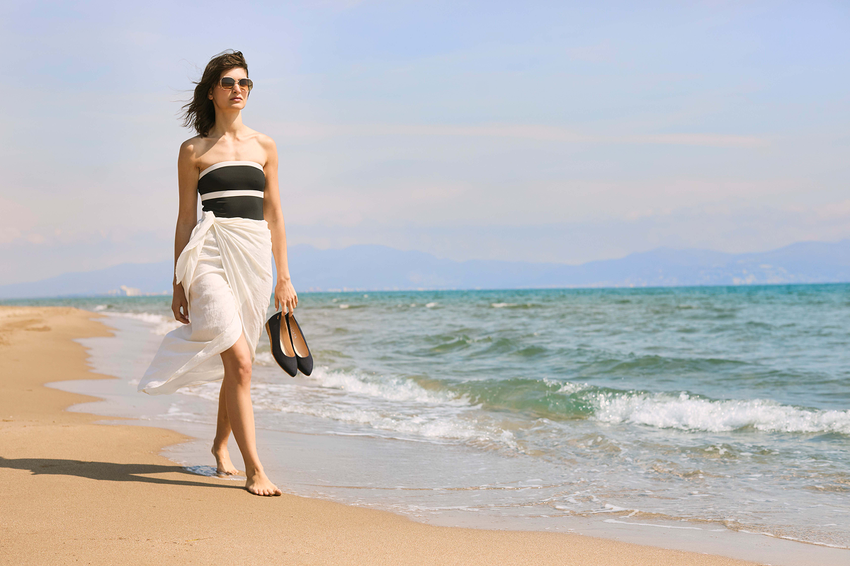 Woman walking barefoot along the shoreline, holding a pair of navy Viscata flats, wearing a black-and-white strapless swimsuit with a white beach wrap beside the sea.