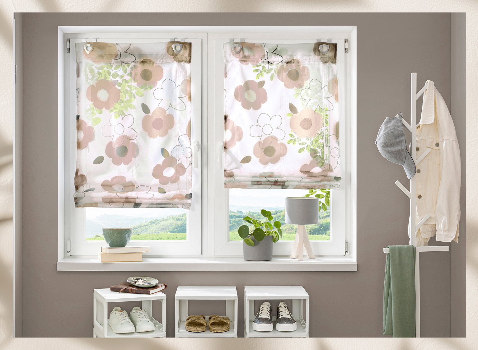 Zwei weiße Rollo-Vorhänge mit buntem Blumenmuster hängen vor einem Fenster, darunter ein Fensterbrett mit Pflanzen, Büchern und einer Lampe