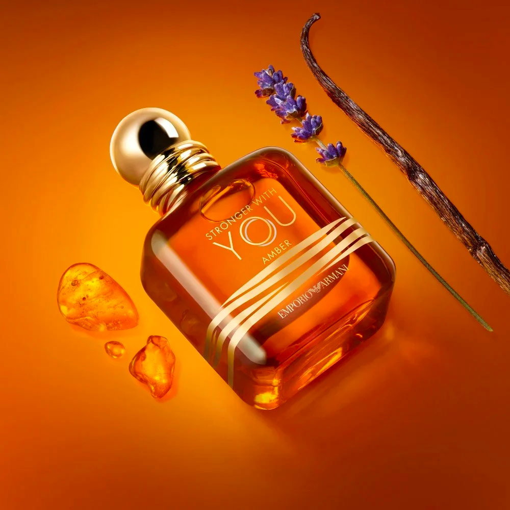 El frasco de la fragancia Armani Stronger With You Amber colocado con varitas de vainilla y piedras de ámbar sobre un fondo naranja brillante.