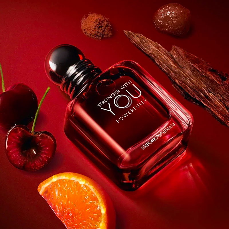 Frasco de Emporio Armani Stronger With You Powerfully presentado sobre una superficie rojo intenso, rodeado de ingredientes inspirados en la fragancia.