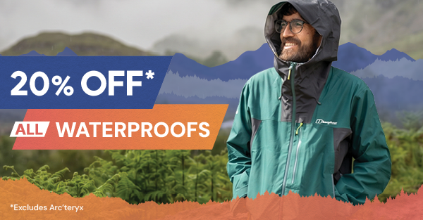 20% OFF ALL WATERPROOFS* *Excludes Arc'teryx. A smiling man walks through a forest, wearing Berghaus Men's Ridge-Seeker GORE-TEX Jacket in Green. 