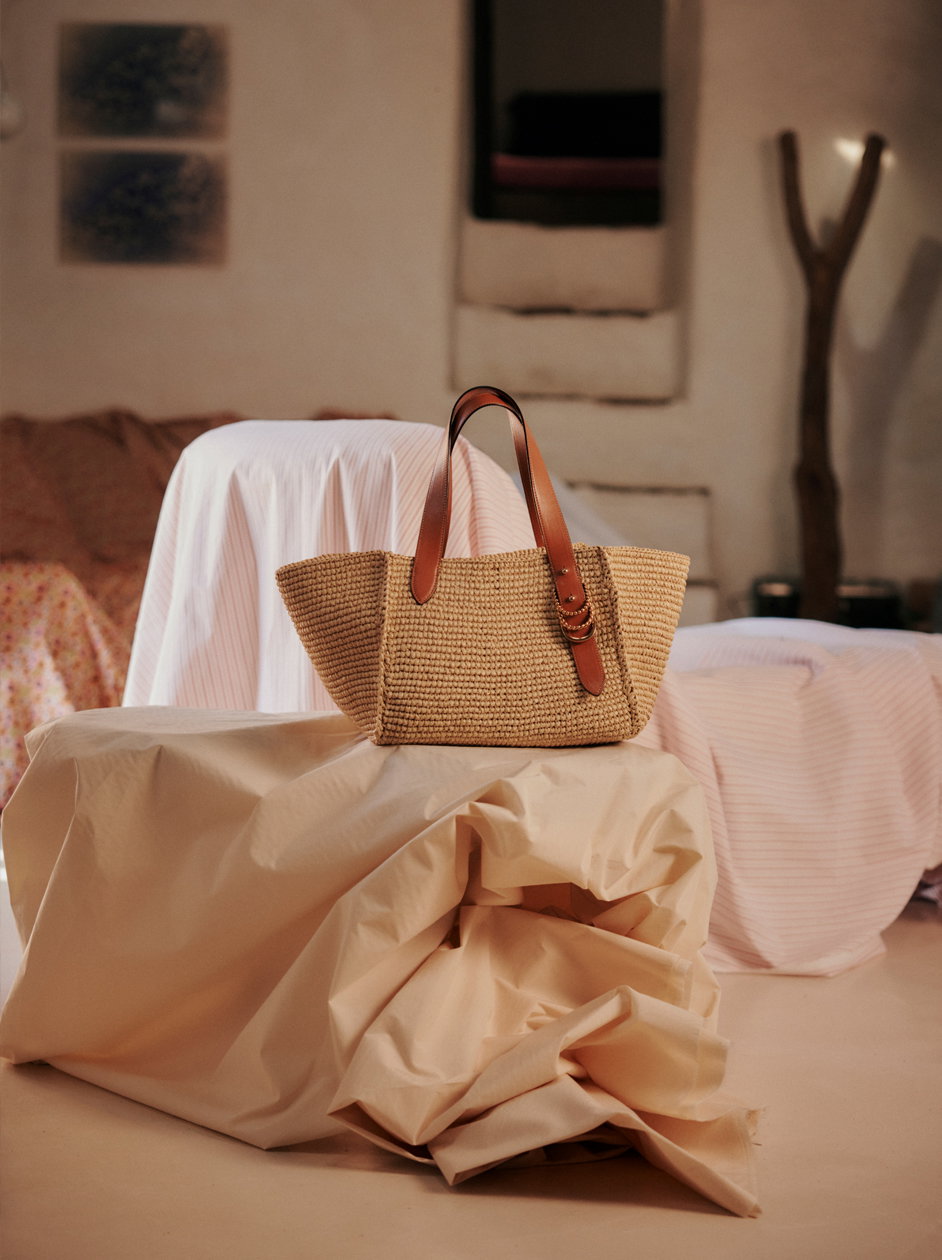 Dorothee Schumacher Strohtasche aus geflochtenem Raffia mit braunen Ledergriffen, inszeniert auf drapiertem Stoff in einem warmen, wohnlichen Interieur mit weichem Licht.